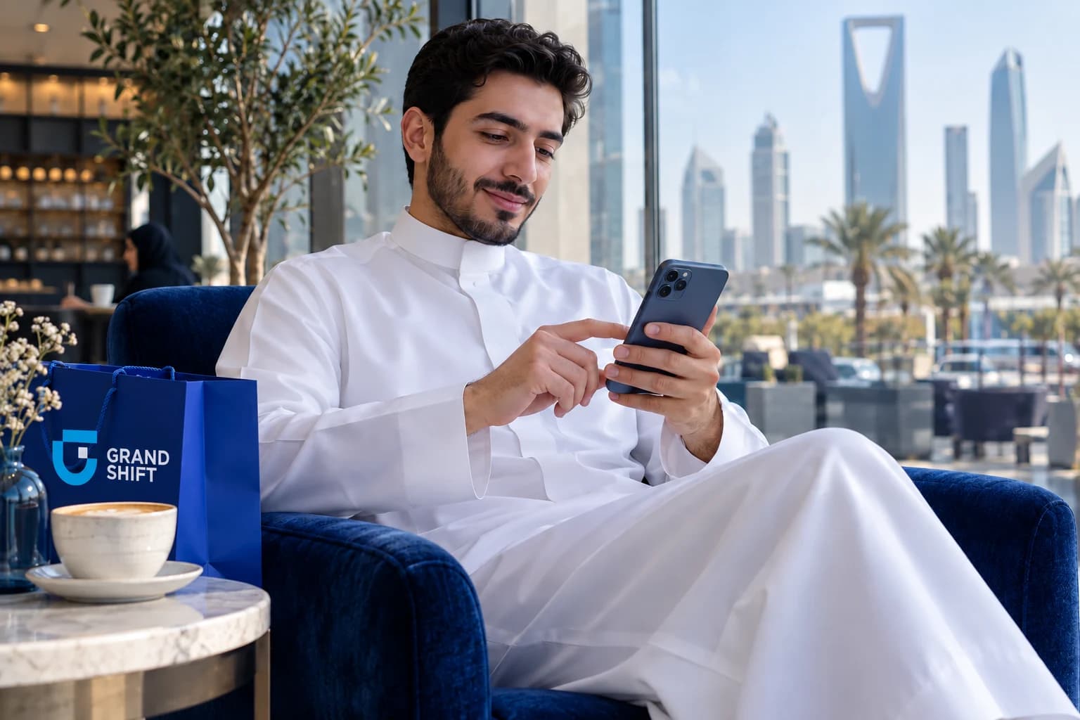 متسوّق سعودي يستخدم هاتفه في مقهى وبجانبه حقيبة تسوّق زرقاء.